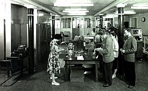 Group of people in a room, gathered around a table with paper tape equipment.
