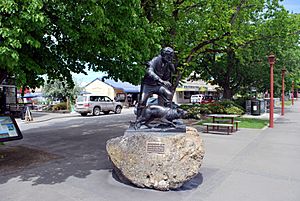 A cast sculpture mounted on a boulder. The sculpture depicts a long haired and bearded man, dressed in a shirt and trousers and carrying a staff. By his side is a sheep dog. Both the man and the dog are facing right. A brass plaque is on the boulder reading "James MacKenzie & dog:Statue by Sam Mahon:Unveiled 7 November 2003". Behind the statue is a paved and treed area with small square patches of grass. A small service road is to the left of the statue, flanked by parked cars and shops.