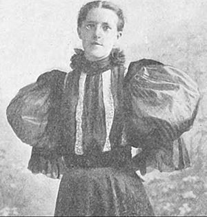 Photograph of a bespectacled woman with her hair pulled away from her face, wearing a frilled high-necked blouse with extremely large puffed sleeves and a striped bodice.