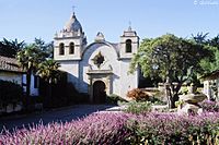 Adobe church with rounded facade and small bell towers on both sides with purple flowers in foreground