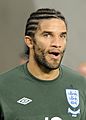 The head and shoulders of a man, with a cornrow hairstyle. On his shirt is a blue and white crest, featuring three lions.