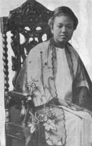 A young Burmese woman seated in a carved wooden chair; she is wearing an embroidered robe over a white dress.