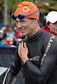 A man wears a black long-sleeved wetsuit, an orange swimming cap, and blue-shaded swimming goggles. He is smiling and holds his left hand over his chest.
