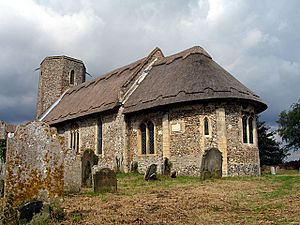 A flint church seen from the southeast, with thatched roofs, an apsidal chancel, a slightly taller nave beyond it, and a tower with an octagonal top