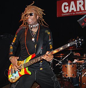 T.M. Stevens wearing black clothing, a variety of jewelry, and black sunglasses, standing onstage in front of a drum kit, playing bass guitar with mouth open