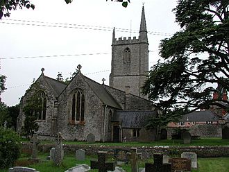 Two bay building with square tower with spirelet. In the foreground are gravestones in a grassy area.