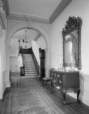 A hallway with rugs stretching through an archway and leading to stairs at end. A console table with drawers sits on the right of the hall with a mirror above it on the wall.
