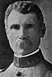 Head of a white man with wavy hair and a bushy mustache wearing a military jacket with the letters "U.S.V." on the high, stiff collar.