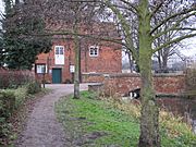 A view of the mill building from the footpath on the north bank.  The pool and the bridge across the race are visible to the right, but the red brick face of the building dominates. In the extreme foreground on the left is a bare-branched tree, its trunk lightly flecked with lichen. In the middle distance on the left is a holly, dark green and dense.