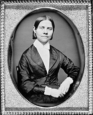 Framed monochrome photograph portrait of a woman sitting, shown from the waist up, left elbow resting on furniture, hands together in lap, the woman wearing a black silk jacket which narrows to conform to the waist, bearing curved lapels, over a plain white blouse with a collar closed at the throat. The woman has dark, straight hair parted in the middle and cut short at the top of the collar. Her head is tilted slightly to her left, face forward, and she is looking directly the observer.