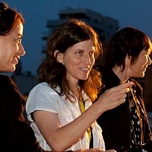 Sperber is shown from the waist up, wearing a white t-shirt. She is standing between two other women, both looking to their left. Sperber is looking to the right of the camera, and pointing. Her hair is reddish, and shoulder-length.