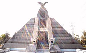 Picture of a Mesoamerican-like pyramid. It is three-stories high. On top, an eagle spreads its wings; at the base, on each side of the central staircase, there is a serpent head.