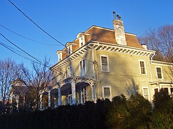A light-colored house with an ornate roofline and curved low roof at the right, with a row of bare trees between it and a distant white house to the left