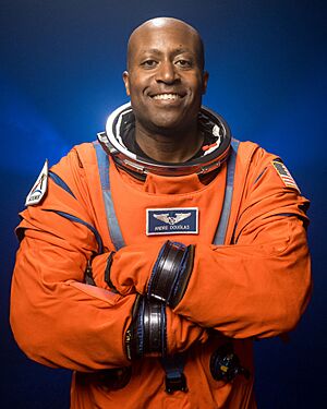 An African American man in an orange flight suit is standing in front of a dark background; he is facing the camera and smiling.