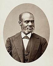 Black and white photo of a Black man of steady countenance and with short, receding hair, and white mutton chops. He is wearing a black suit jacket and vest over a white shirt.