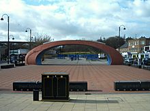 A brick-red, elliptically curved arch, twice as wide as it is high, over an open area with a brick-red surface