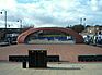 A brick-red, elliptically curved arch, twice as wide as it is high, over an open area with a brick-red surface