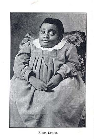 A photographic portrait from 1906 of a small African-American woman, seated, hands folded in lap. She is wearing a voluminous long-sleeved dress with a white collar.