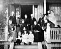 Late 19th century photo of a family of ten adults and four children posing on the front porch of a large house. The women wear long dresses with high collars and leg-o-mutton sleeves. The men have flowing moustaches. Marjory is a toddler in a long dress and black stockings.