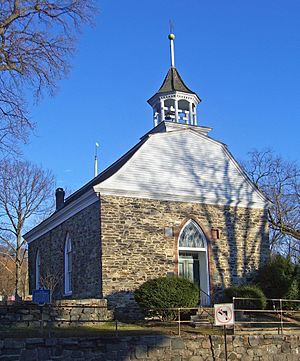 A small stone building with a bell-shaped roof and upper section sided in wood seen from slightly below and to its right. There is a wooden bell tower on top with a weathervane. All the windows have rounded tops that end in points.