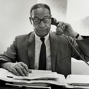 Ted Berry is seen wearing a suit and tie with thick-rimmed glasses, speaking on the telephone, with a pencil in hand, glancing down at a stack of various papers
