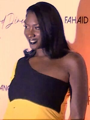 blurry image of Aya Nakamura wearing a black-and-yellow fitting dress that reveals left shoulder, standing onstage, grinning and looking right of camera