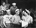 A military scene.  A woman dressed as a nurse holds a skirt she has just received from an enlisted man; she is pleased while he appears self-deprecating. Other enlisted men, many bare-chested, watch.