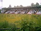 A grandstand whose coloured seats spell out the word Darlington. A floodlight pylon stands at one end. The stand faces a grassed area overgrown with weeds.