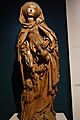 A wooded sculpture of St. Elizabeth. She has on a 15th-century style dress and wears a cloak which falls to mid shin. She holds a pitcher in her left hand and at her right foot a small depiction of a man kneels and gazes up at her.