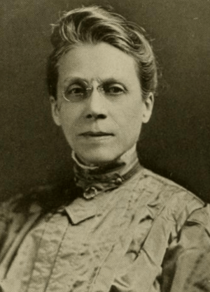 A white woman wearing pince-nez glasses and a high-collared dress with pleated front; her hair is dressed back from her face and up at the nape