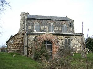A stone church with a brick porch seen from the south.  To the left are the remains of the collapsed tower.  The clerestory contains three windows and six carved panels.