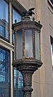 A decorative bronze lamppost with narrow panes of clouded glass in a circular pattern. Behind it is a window in a stone building.