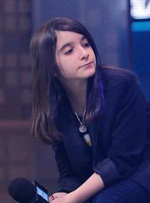 A white teenager with long brown hair is sitting, holding a microphone, wearing a dark blue shirt.  She is leaning to her right, looking to her left—right of the camera.