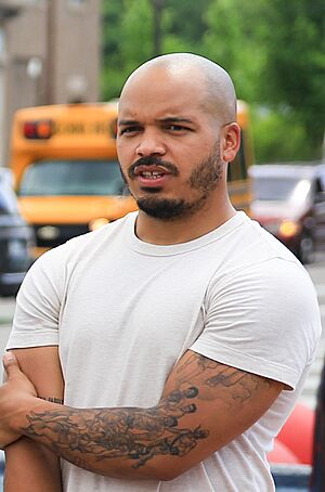 A young, black man with a mustache and anchor beard sits behind a laptop and stares off to his left. Another person, barely visible, is seated to Ellison's right.