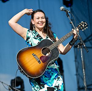 Leela Gilday wearing a patterned dress, standing onstage in front of a microphone with a guitar, squinting and smiling ahead, adjusting sunglasses on top of her head with her right hand.