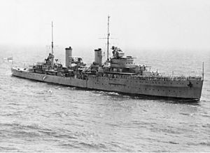 A two funnelled cruiser at sea. People are clustered on the forward deck