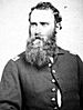 Head and torso of a white man with a long, full beard and curly hair, wearing a military jacket, unbuttoned at the stomach, over a white shirt.