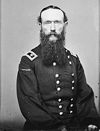Black and white photo shows a heavily-bearded man staring directly into the camera. He wears a dark, double-breasted military uniform with general's stars on the shoulder tabs.