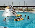 The Apollo spacecraft and orange rafts float in a pool, surrounded by divers