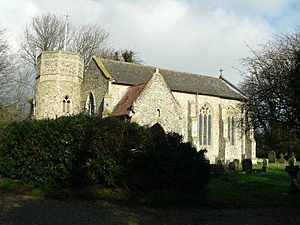 A flint church seen from the southwest.  From the left are a round tower with an octagonal top, the west gable, the south porch, and the south wall of the nave with large windows