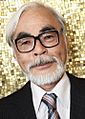 Close up photograph of Hayao Miyazaki, smiling and wearing a suit and tie in front of a gold-colored mosaic.