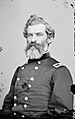Head and torso of a white man with thick wavy hair and a full beard, wearing a double-breasted military jacket with a rectangular patch on each shoulder.