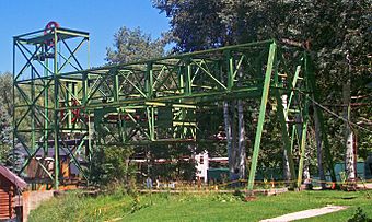 An L-shaped green open latticework steel structure on a small rise. The long end runs parallel to the ground to where it is supported by two diagonal beams in front of some deciduous trees with light trunks. On the bottom of the middle section is another section housing a large red pulley wheel parallel to the ground. A smaller red pulley and cable can be seen in the top of the short section.
