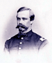 Head and shoulders of a white man with a bushy Van Dyke mustache, wearing a jacket with buttons down the center and rectangular patches on each shoulder.