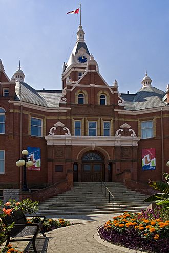 A blue sky is the background for a red brick building extending past the frame of the photograph to either side. At bottom right is a portion of an arcing flowerbed, populated with orange flowers in full bloom, to their left a curving walkway and a decorative iron bench. The pathway leads to stairs, wide at the bottom and narrowing to the entrance doors, with a central iron railing and bounded at each side by a short brick wall, from which base protrudes a black iron pole with four visible light globes. Above the door are three framed windows, above them two more inset at the end of a cross gable, on each side of which is visible the main structure's roof sloping towards the viewer. Behind the gable rises a tower, atop which is a clock, all surmounted by a pole from which flies the Canadian flag.