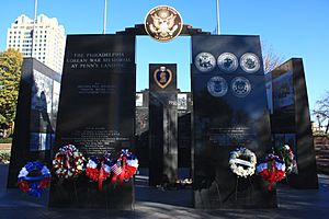 The memorial's western facade is shown, composed of two 16-foot-tall black granite monoliths with inscriptions. Veterans Day wreaths are on stands in front of the monoliths. Several more black granite monoliths are beyond and to the sides of the facade, arranged in a square pattern. A single black granite column is visible in the middle of the square with the year 1950 at the top and the inscribed names of those killed or missing in action are listed below.