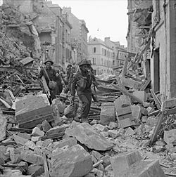 A line of soldiers clambering over the heaped rubble of destroyed buildings in a badly-damaged street.