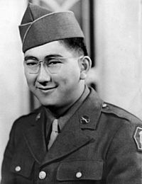 Head and shoulders of a smiling young man with dimples and round wire-framed glasses wearing a garrison cap and a military jacket over a shirt and tie