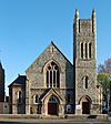 A pale stone church on a corner site, with a dominant square tower with spirelets. To the left of this is the entrance, below a four-light lancet window and three sexfoils.  To the right is the main body of the church, with two side chapels and lancet windows.