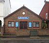 A small, low hall in two colours of brick, with brown-framed windows and a matching double door flanked by blue posters. Above this is a blue panel with "THE WORTHING CHRISTADELPHINS" and a round window.